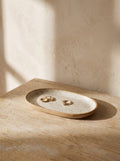 Oval speckled oatmeal stoneware trinket tray with gold earrings on an oak dressing table