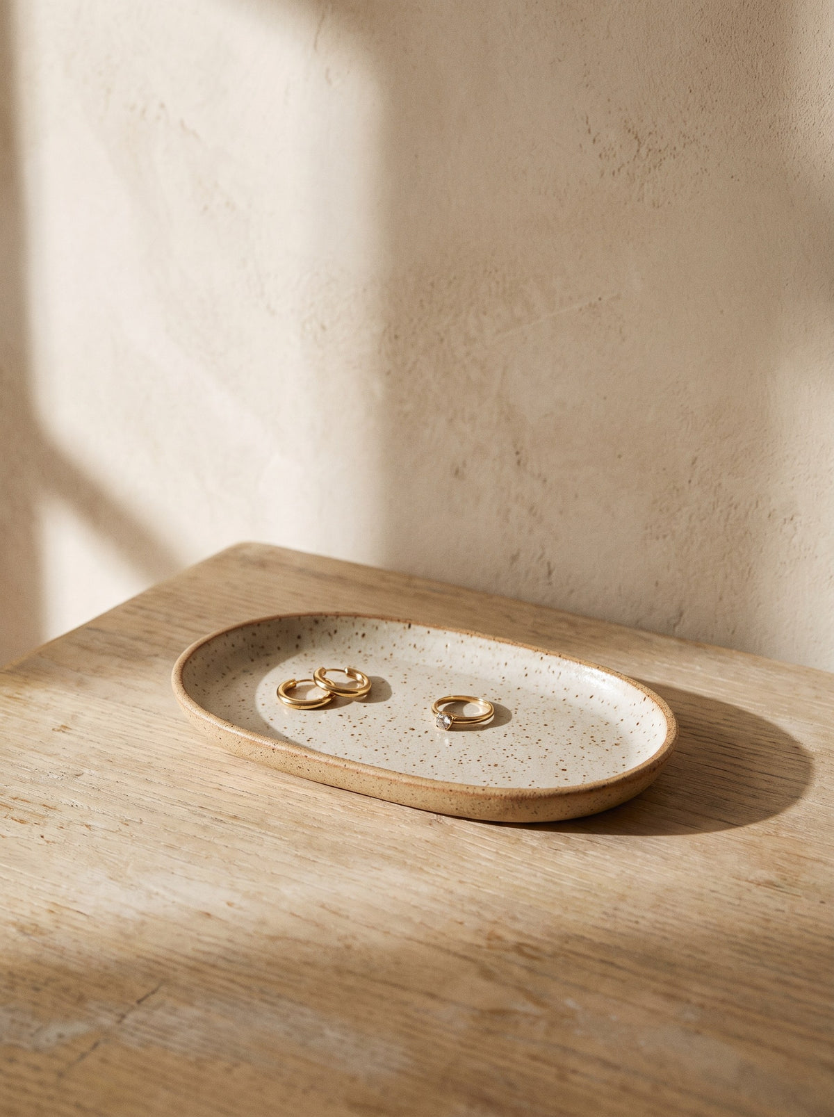 Oval speckled oatmeal stoneware trinket tray with gold earrings on an oak dressing table