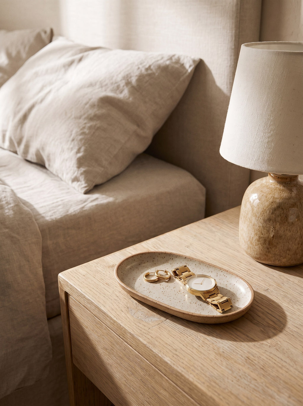 Speckled stoneware trinket tray on a bedside table holding gold hoop earrings and a watch