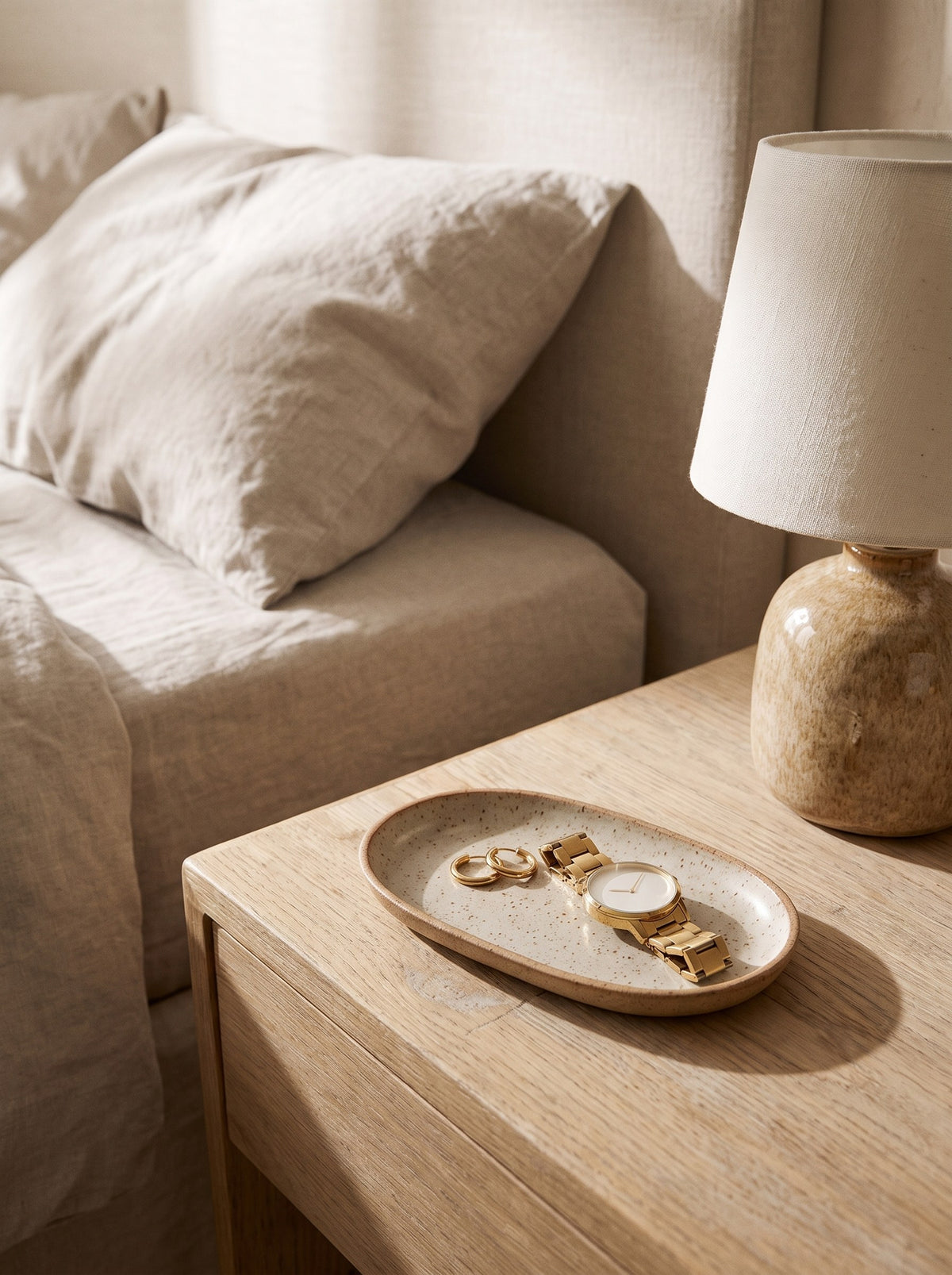 Speckled stoneware trinket tray on a bedside table holding gold hoop earrings and a watch